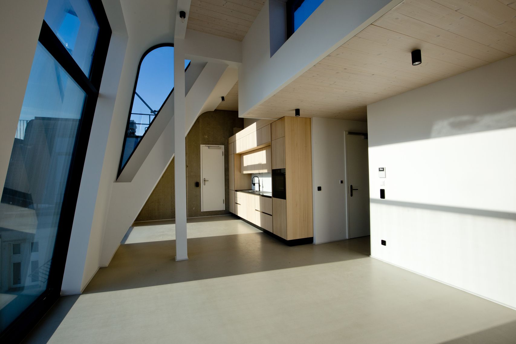 Moderner offener Wohnraum mit großer Fensterfront, sichtbaren Holzbalken und minimalistischer Einbauküche aus hellem Holz