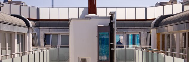 Blick über eine Dachterrasse mit Glasgeländern und weißen Gebäudeteilen unter blauem Himmel