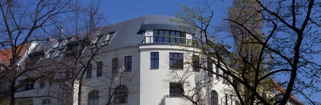 Mehrstöckiges weißes Gebäude mit abgerundetem Eckbereich und mehreren Fenstern, umgeben von kahlen und belaubten Bäumen