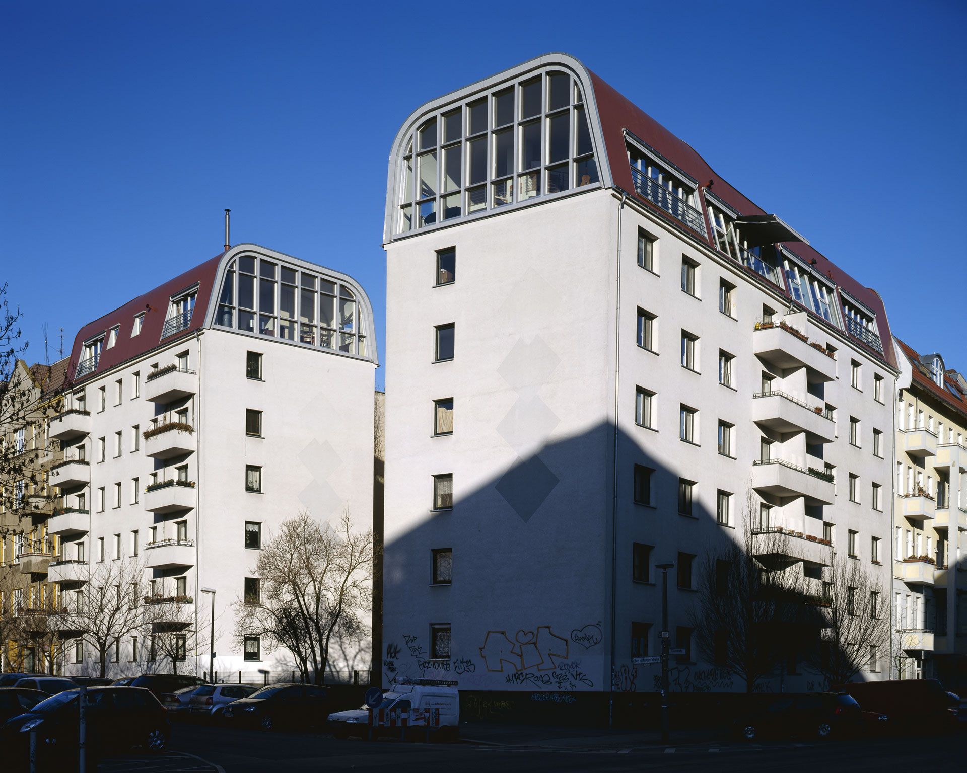 Zwei mehrstöckige Wohngebäude mit weißen Fassaden und roten Dachaufbauten vor blauem Himmel