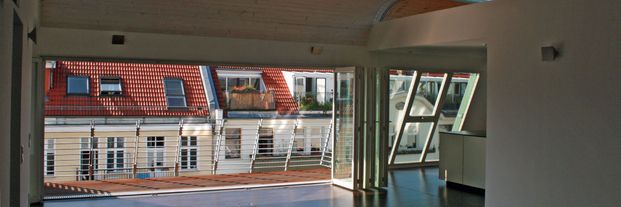Großer leerer Wohnraum mit offenem Fenster und Blick auf rote Dachziegel und weiße Fassaden gegenüberliegender Gebäude